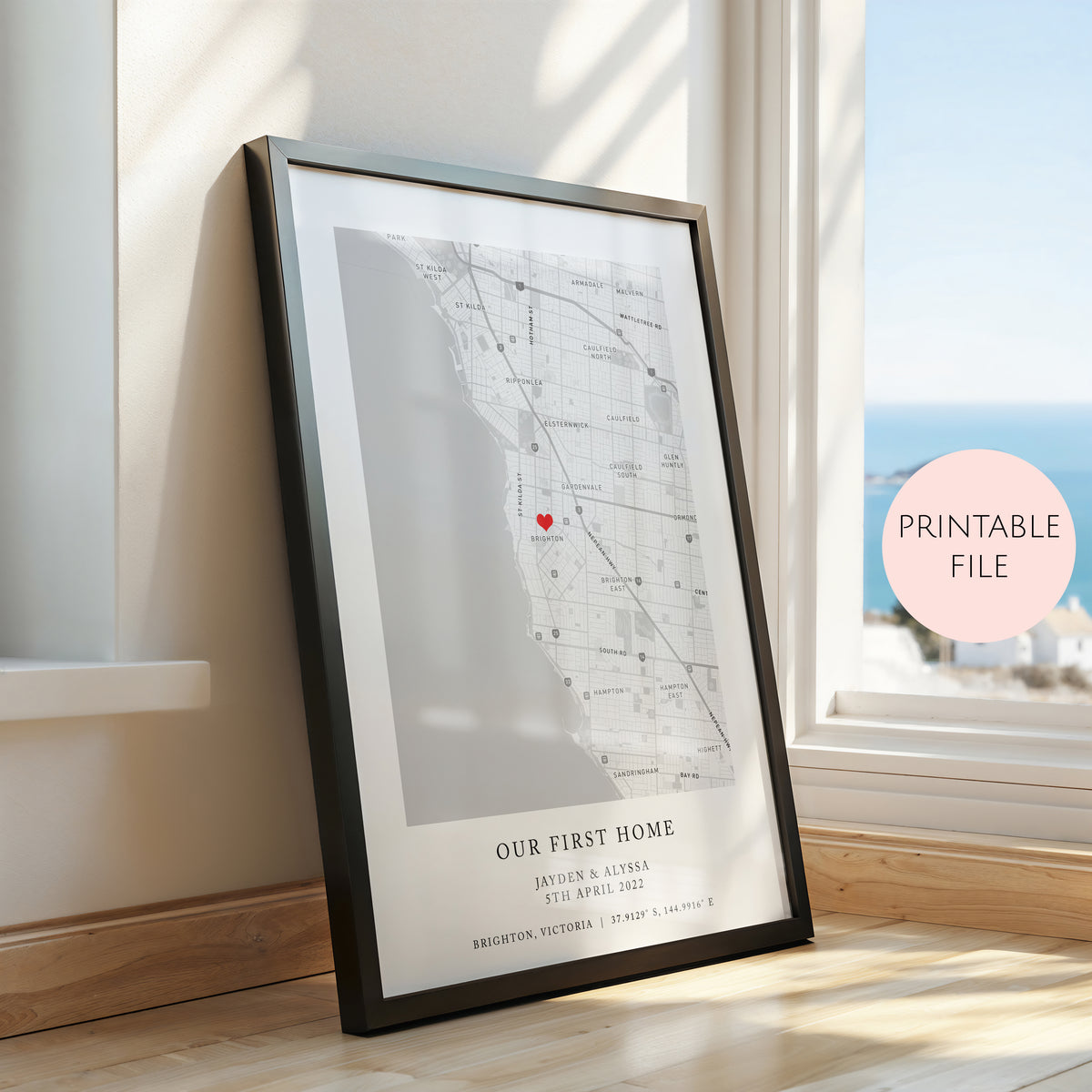 A framed personalised map print leaning against a window on a wooden floor. The print shows a street map of Brighton, Victoria with a small red heart marking a location, plus the text 'OUR FIRST HOME' and custom names/date.