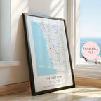 A framed personalised map print leaning against a window on a wooden floor. The print shows a coastal street map with the text 'OUR FIRST HOME' and names/date/location printed at the bottom.