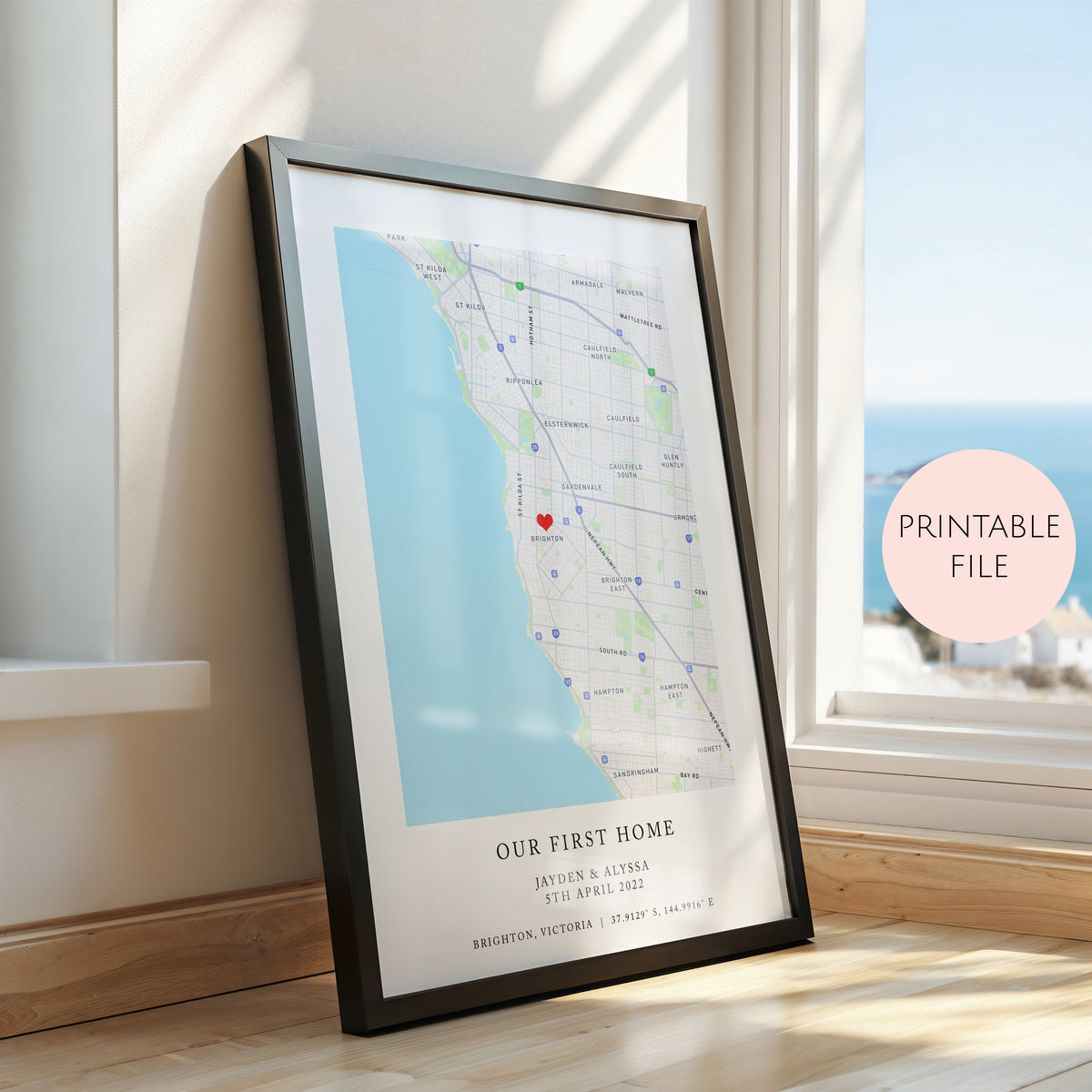 A framed personalised map print leaning against a window on a wooden floor. The print shows a coastal street map with the text 'OUR FIRST HOME' and names/date/location printed at the bottom.