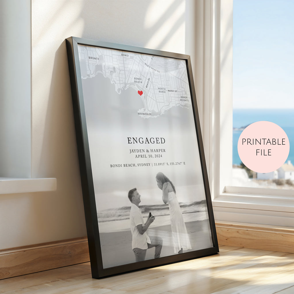 A framed personalised engagement print leaning against a window on a wooden floor. The print features a black and white map with a small red heart, the word 'ENGAGED,' couple names and date, and a black and white photo of a couple on a beach.