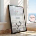 A framed personalised engagement print leaning against a window on a wooden floor. The print features a black and white map with a small red heart, the word 'ENGAGED,' couple names and date, and a black and white photo of a couple on a beach.
