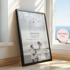 A framed personalised engagement print leaning against a window on a wooden floor. The print features a black and white map with a small red heart, the word 'ENGAGED,' couple names and date, and a black and white photo of a couple on a beach.