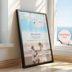 A framed personalised map and photo print leaning against a window on a wooden floor. The print includes a beach map, the word 'ENGAGED,' names and date of a couple, coordinates, and a faded photo of the couple on the beach.