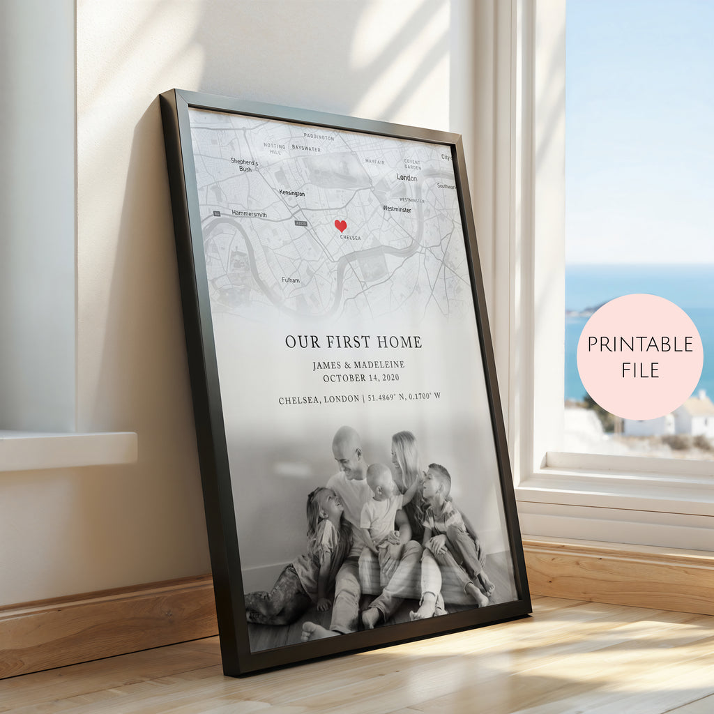A framed personalised map print leaning against a window in a sunlit room. The print shows a black and white city map with a small red heart, the text 'OUR FIRST HOME' and custom names/date/location, and a black-framed photo of children sitting on the floor.