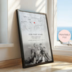 A framed personalised map print leaning against a window in a sunlit room. The print shows a black and white city map with a small red heart, the text 'OUR FIRST HOME' and custom names/date/location, and a black-framed photo of children sitting on the floor.