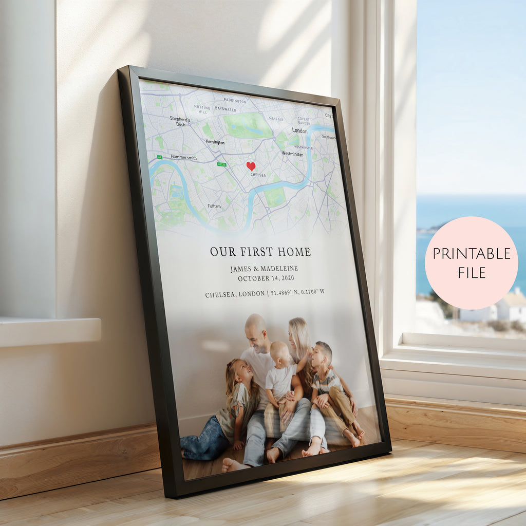 A framed personalised map print leaning against a window on a wooden floor. The black framed poster shows a map with a red heart marker, the text 'OUR FIRST HOME' and custom names/date/location, and a photo of children sitting on the floor.