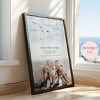 A framed personalised map print leaning against a window on a wooden floor. The black framed poster shows a map with a red heart marker, the text 'OUR FIRST HOME' and custom names/date/location, and a photo of children sitting on the floor.