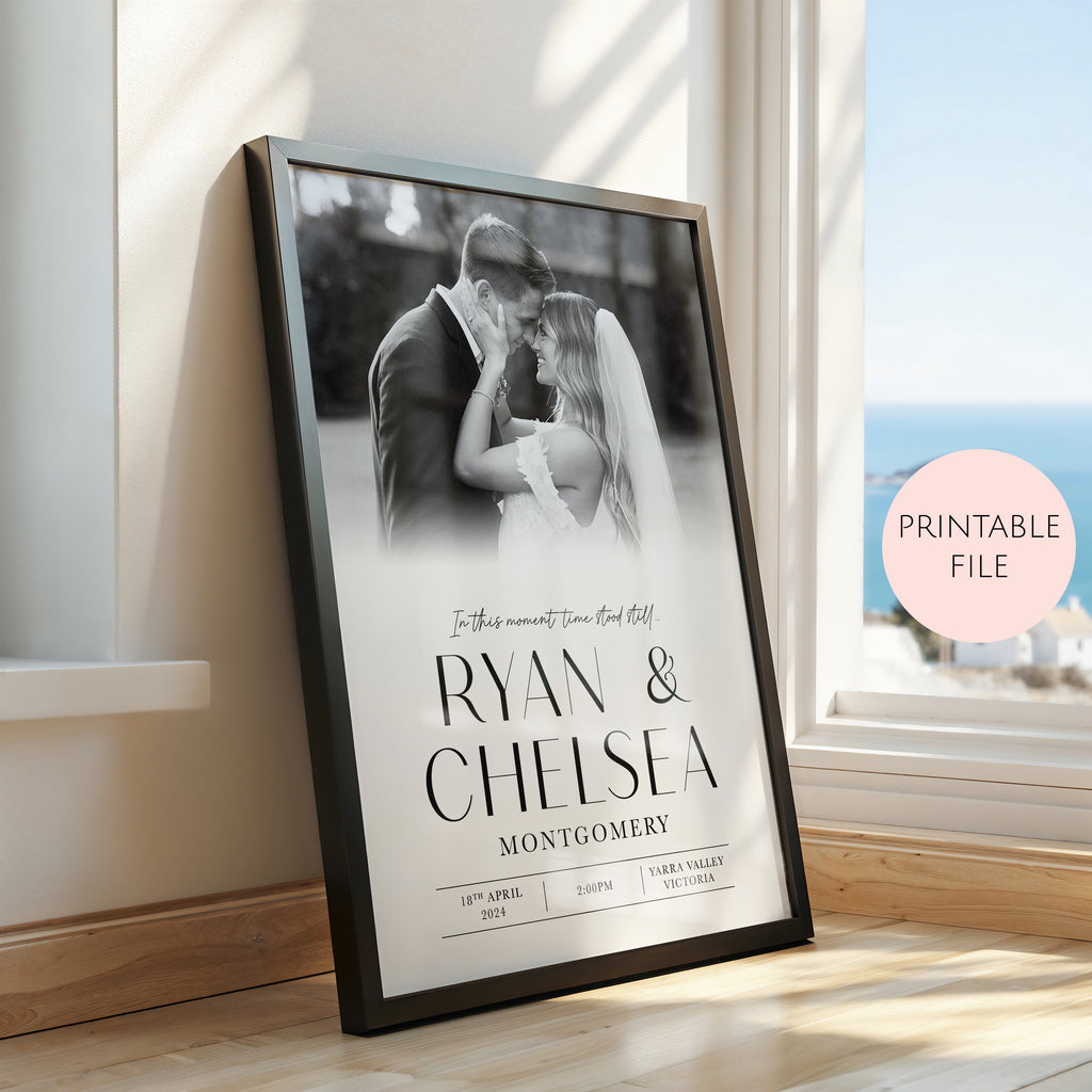 A framed personalised wedding photo print leaning against a window on a wooden floor. The black frame contains a black and white photo of a couple with the names 'RYAN & CHELSEA' and 'MONTGOMERY' printed in large black text, along with wedding date details.