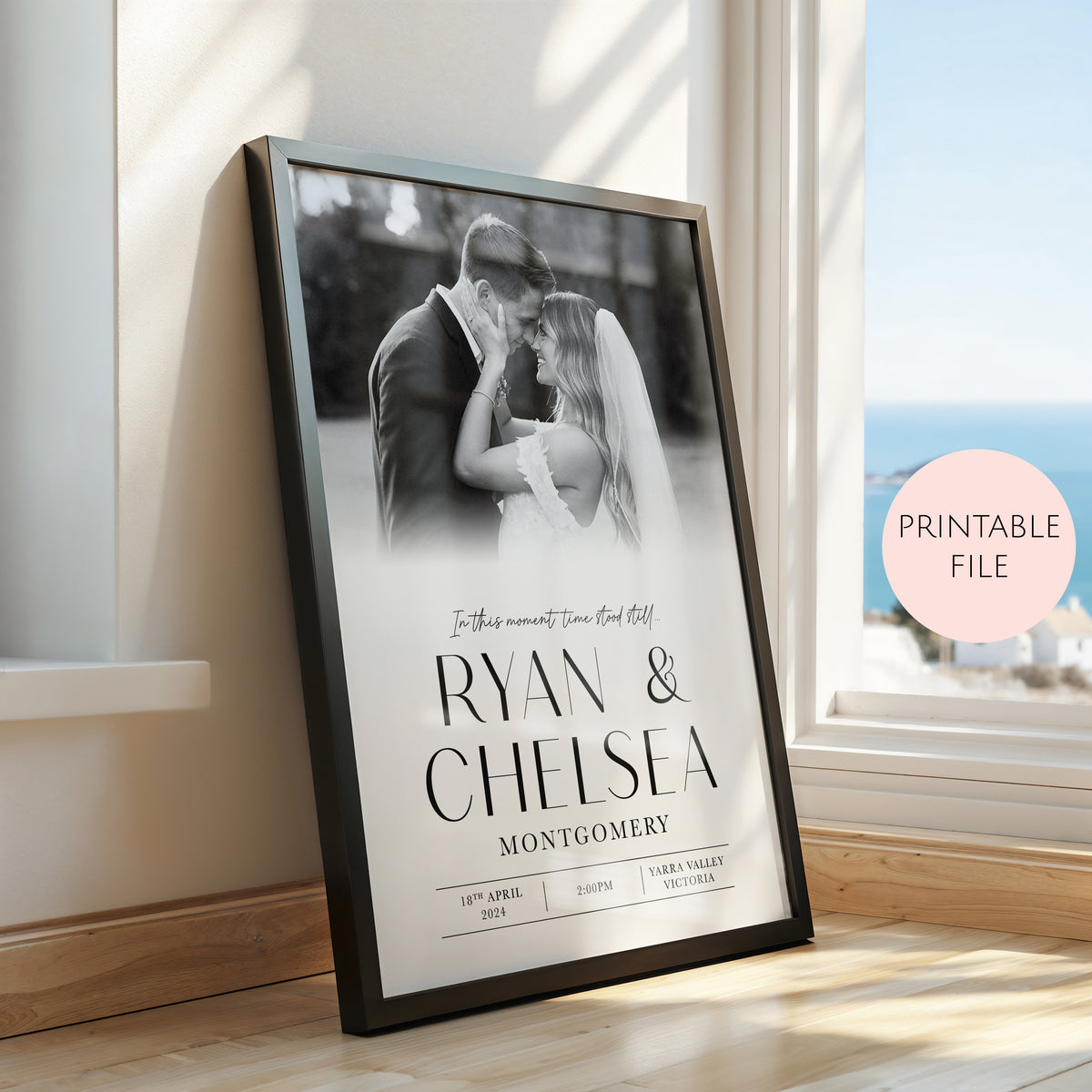 A framed personalised wedding photo print leaning against a window on a wooden floor. The black frame contains a black and white photo of a couple with the names 'RYAN & CHELSEA' and 'MONTGOMERY' printed in large black text, along with wedding date details.