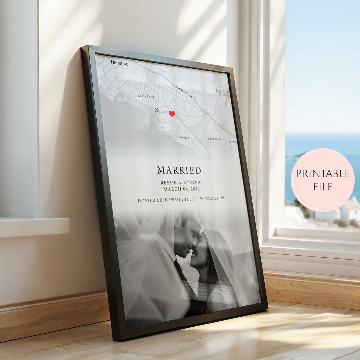 A framed custom map and photo print leaning against a window on a wooden floor. The print features a black framed minimalist design with a map, a small red heart, the word 'MARRIED,' couple names and date, and a soft black and white wedding photo.