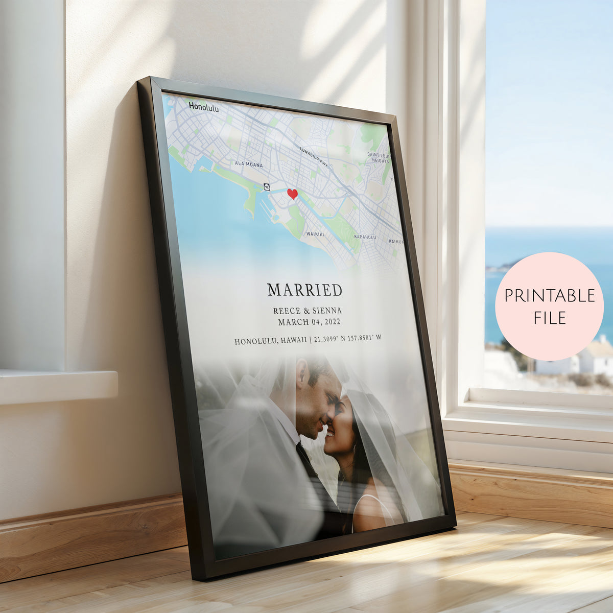 A framed personalised map and photo print leaning against a window on a wooden floor. The black frame contains a map of Honolulu with a red heart marker, the text 'MARRIED', names and date, and a faded couple photo.