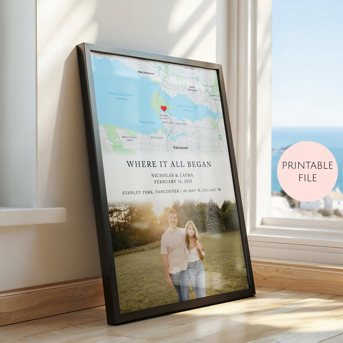 A framed personalised map and photo print leaning against a window in a sunlit room. The print includes a light blue and green map with a red heart marker, text reading 'WHERE IT ALL BEGAN' and names/date, plus a couple's photo at the bottom.