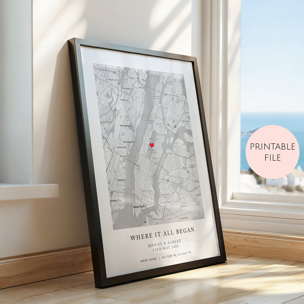 A framed personalised map print leaning against a window on a wooden floor. The print features a black and white street map with a small red heart near the centre and the text 'WHERE IT ALL BEGAN' with names and date below.