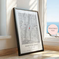 A framed personalised map print leaning against a window on a wooden floor. The print features a black and white street map with a small red heart near the centre and the text 'WHERE IT ALL BEGAN' with names and date below.
