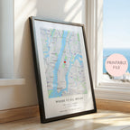 A framed custom map print of New York City leaning against a window on a wooden floor. The print features a light blue and gray map with a small red heart near the center, black text at the bottom reading 'WHERE IT ALL BEGAN', and a black frame.