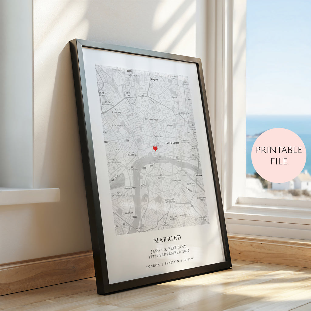 A framed personalised map print leaning against a wall on a wooden floor next to a window. The print shows a street map with a small red heart marker and the text 'MARRIED' and names/date below.