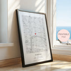 A framed personalised map print leaning against a wall on a wooden floor next to a window. The print shows a street map with a small red heart marker and the text 'MARRIED' and names/date below.
