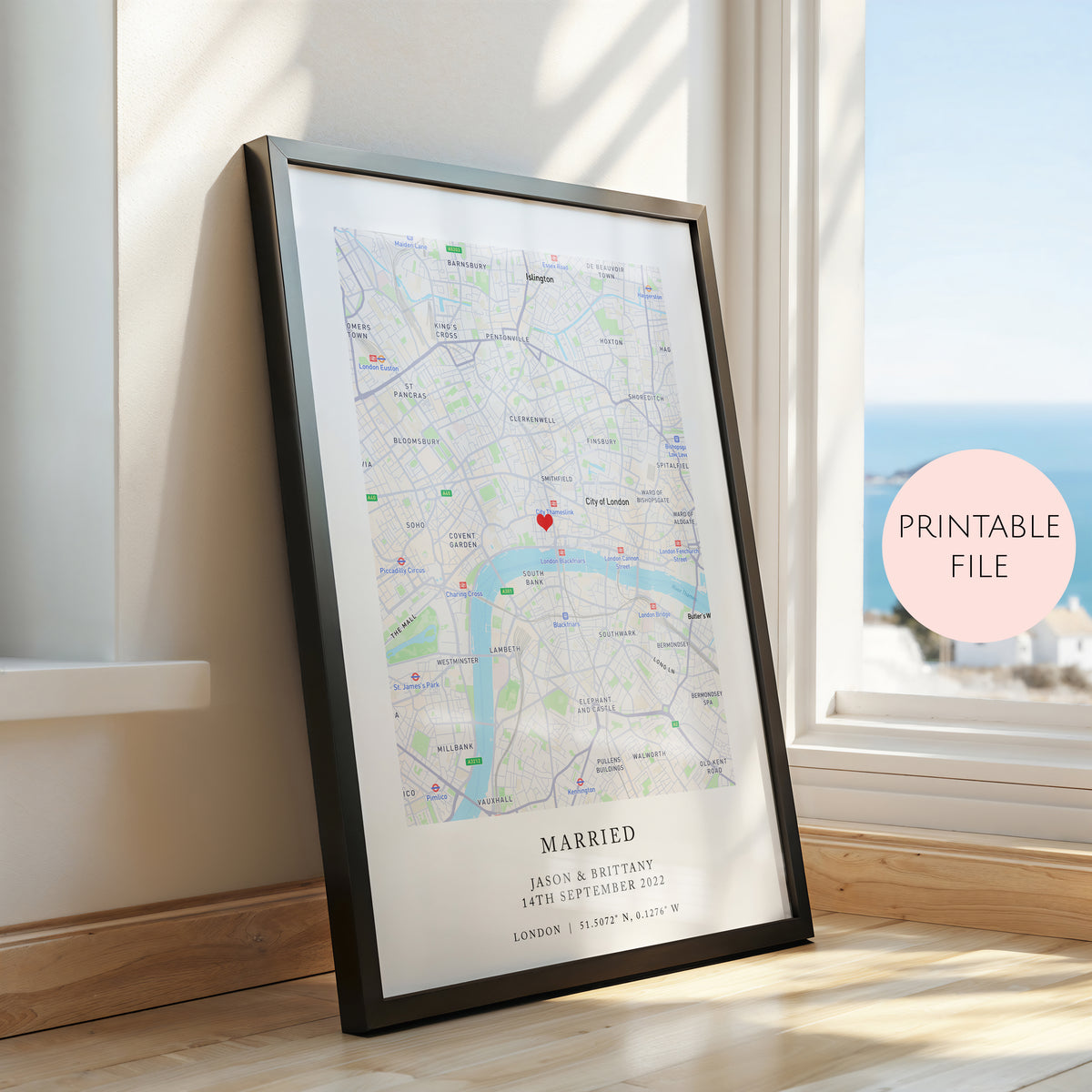 A framed personalised map print of London leaning against a window on a wooden floor. The black framed print features a detailed map with a red pin marking a location, personalised text reading 'MARRIED' and names/date, and a soft pink circular badge that says 'PRINTABLE FILE'.