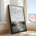 A framed personalised map and photo print leaning against a window on a wooden floor. The print includes a black and white map with a red heart marking, and a black and white couple photograph.