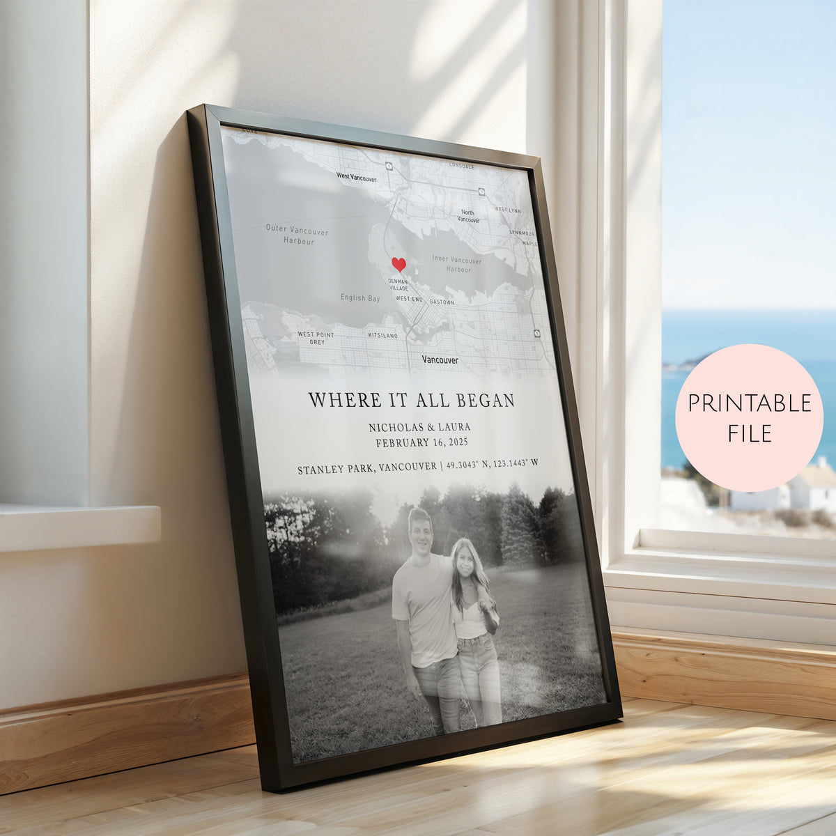 A framed personalised map and photo print leaning against a window on a wooden floor. The print includes a black and white map with a red heart marking, and a black and white couple photograph.